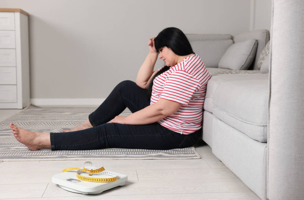 Depressed overweight woman sitting near sofa at home, focus on scales with measuring tape - Foto, afbeelding