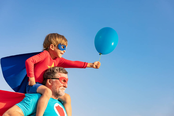 Superhero senior man and child playing outdoor. Super hero grandfather and boy having fun together against blue summer sky background. Family holiday concept. Happy Father's day - Foto, imagen