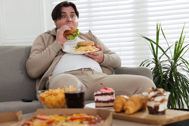 Overweight man with plate of burgers and French fries on sofa at home - Fotografie, Obrázek