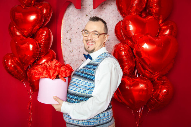 Hombre de estilo retro barbudo divertido en el chaleco azul que sostiene la caja presente en los globos de forma de corazón rojo para el día de San Valentín - Foto, Imagen