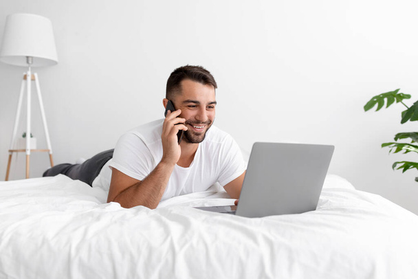 Zadowolony biały atrakcyjny młody mężczyzna w białej koszulce obudził się rozmawiając, surfując, pracując nad komputerem i dzwoniąc - Zdjęcie, obraz