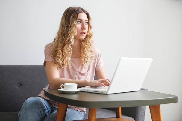 Beautiful young woman sitting on the sofa with white cup of coffee, working on white laptop. Girl in pink shirt looking to the monitor. Distance learning student - Φωτογραφία, εικόνα