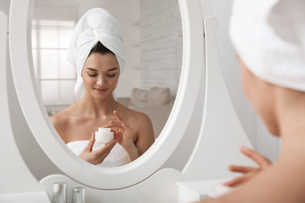 Beautiful young woman with hair wrapped in towel holding face cream near mirror at home - Photo, image