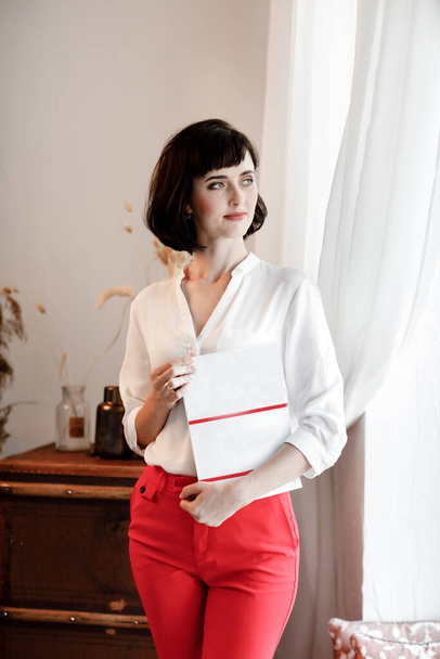 belle jeune femme aux cheveux courts foncés tenant un magazine avec une maquette. fille dans une chemise blanche et pantalon rouge debout près de la fenêtre. - Photo, image