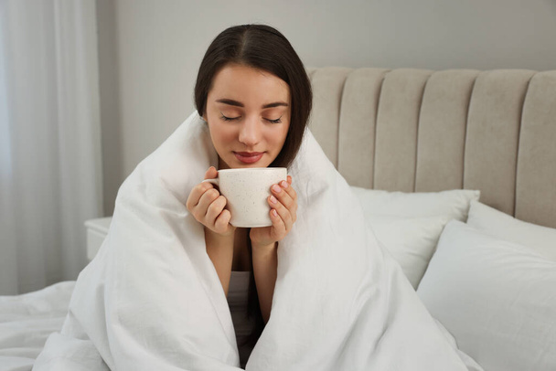 Beautiful young woman with cup of drink wrapped in blanket at home - Foto, afbeelding