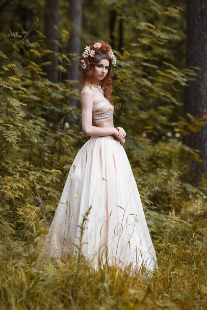 A slender girl in a long light dress with flowers in her hairstyle outside. - Foto, afbeelding