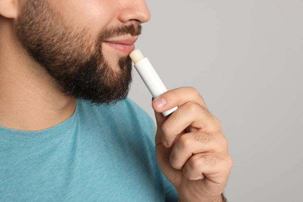 Young man applying lip balm on grey background, closeup - Фото, зображення