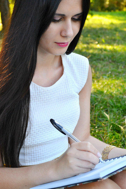 młoda brunetka kobieta z piękne dłonie, pisząc w swoim pamiętniku i uśmiechając się - Zdjęcie, obraz