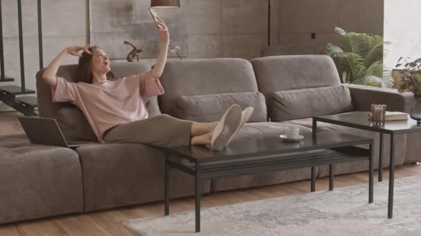 Stab slowmo shot of young Caucasian woman in casualwear taking funny selfie portraits on smartphone while working from home, sitting on sofa with laptop in modern apartment - Footage, Video