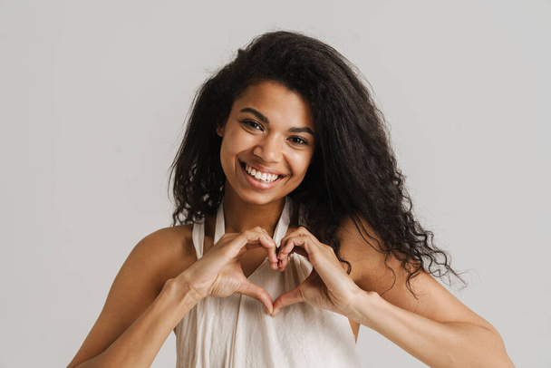 Smiling young african woman in casual wear standing over isolated gray background showing heart gesture - Фото, изображение