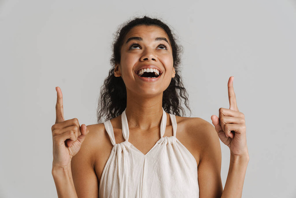Black young woman in dress smiling and pointing fingers upward isolated over white background - Φωτογραφία, εικόνα