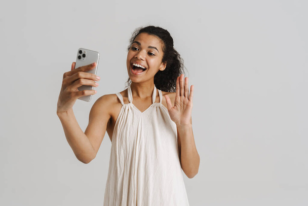 zwart jonge vrouw gebaren terwijl het nemen van selfie op mobiele telefoon geïsoleerd over witte achtergrond - Foto, afbeelding