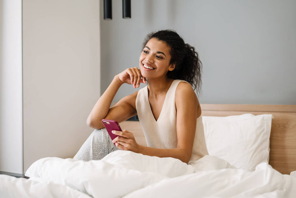 Happy young african woman sitting in bed in the bedroom holding mobile phone - Foto, Bild