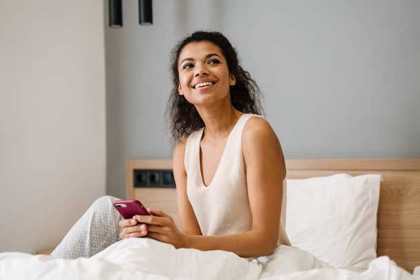 Happy young african woman sitting in bed in the bedroom holding mobile phone - 写真・画像