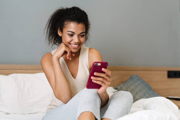 Happy young african woman sitting in bed in the bedroom holding mobile phone - Foto, afbeelding