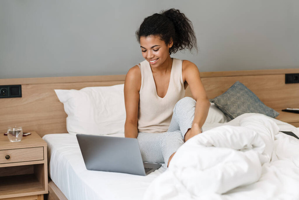Young black woman using laptop while sitting on bed after sleep at home - Foto, Imagen