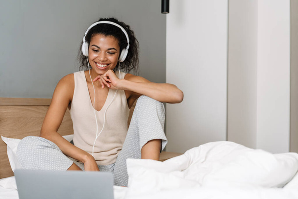 Happy young african woman sitting in bed in the bedroom wearing earphones talking on a video call via laptop computer - 写真・画像
