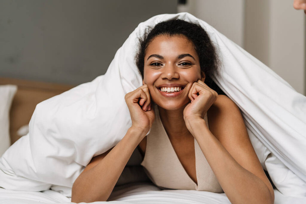 Happy young african woman laying hiding under a blanket on a bed at home laughing - Foto, immagini