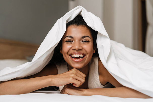 Happy young african woman laying hiding under a blanket on a bed at home laughing - Foto, Bild