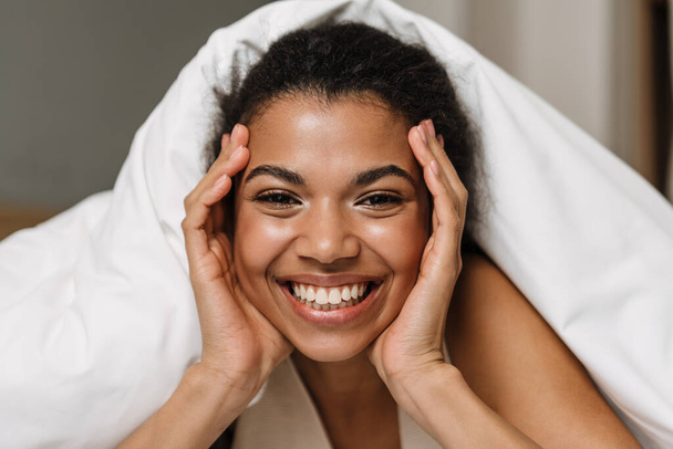 Happy young african woman laying hiding under a blanket on a bed at home laughing - Photo, Image