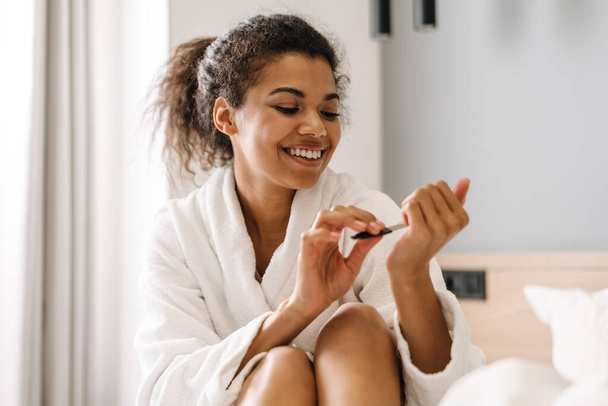 Smiling young african woman in bathrobe sitting on a bed filing her nails in the bedroom - Φωτογραφία, εικόνα
