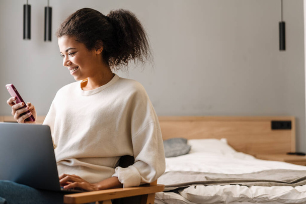 Smiling young african woman using laptop computer while sitting on a chair in a bedroom holding mobile phone - Photo, image