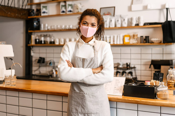 Young black waitress in face mask standing by cafe counter indoors - Fotografie, Obrázek