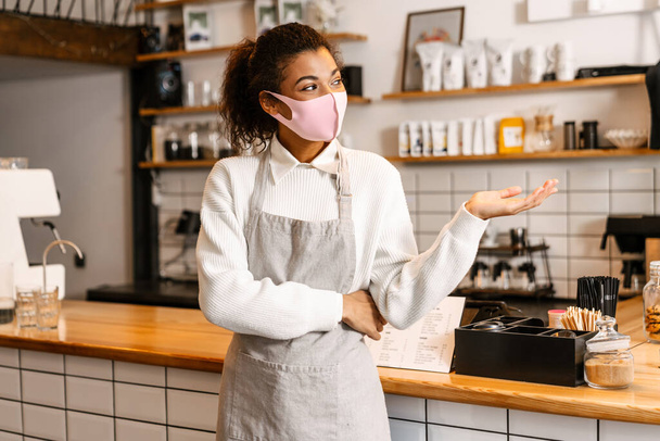 Young black waitress in face mask holding copyspace while standing on workplace indoors - Фото, зображення