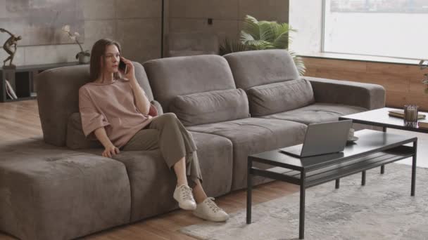 Slow-Motion-Aufnahme einer jungen Kaukasierin, die auf dem Sofa in einer modernen Wohnung mit dem Handy spricht - Filmmaterial, Video
