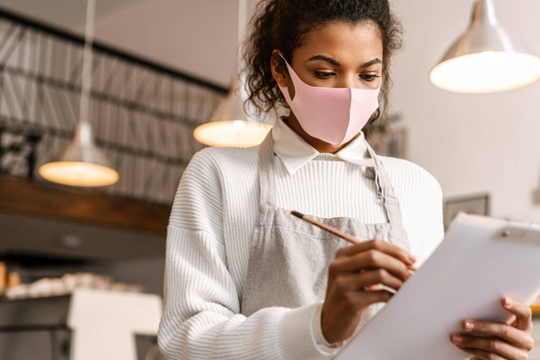 Young black waitress wearing face mask writing down notes on clipboard in cafe - Foto, immagini