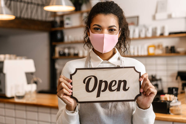 Young black waitress in face mask showing open sign board at cafe indoors - Photo, image