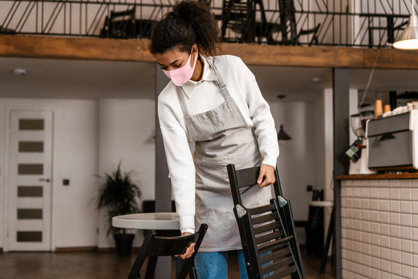Young black waitress wearing face mask and apron working in cafe indoors - Fotó, kép