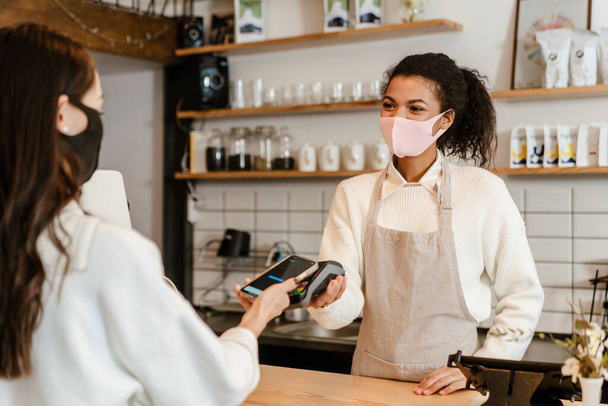 Young black waitress in face mask working with payment terminal at cafe counter indoors - Photo, Image