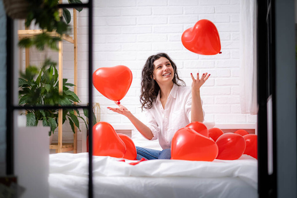 Brunette European girl rejoices playing on bed with red heart shape balloons. Morning love surprise gift on valentines day - Foto, Imagem