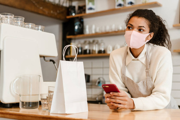 Young black waitress wearing face mask using cellphone while working in cafe indoors - Foto, imagen