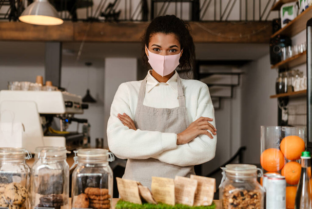 Black woman barista in face mask standing by cafe counter indoors - Photo, Image