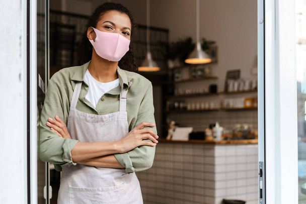 Young black waitress wearing face mask standing in doorway at cafe - Foto, Imagem