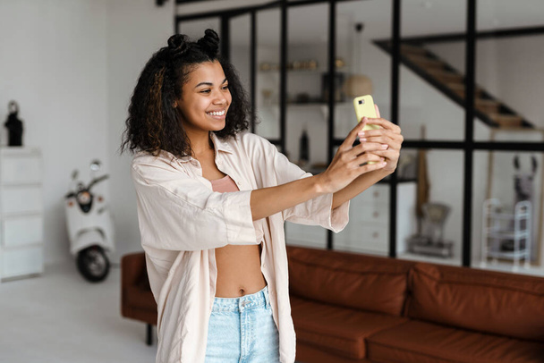 Jeune femme noire souriant tout en prenant selfie sur téléphone portable à la maison - Photo, image