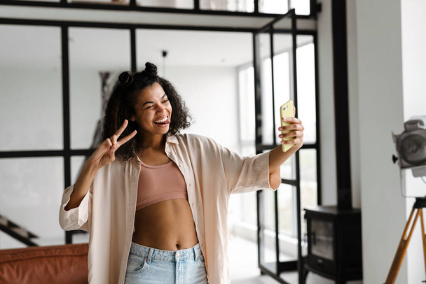 Jeune femme heureuse afro-américaine tenant un téléphone intelligent faisant un vlog de la maison - Photo, image