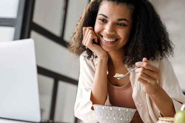 Black young woman working with laptop while having breakfast at home - Photo, image