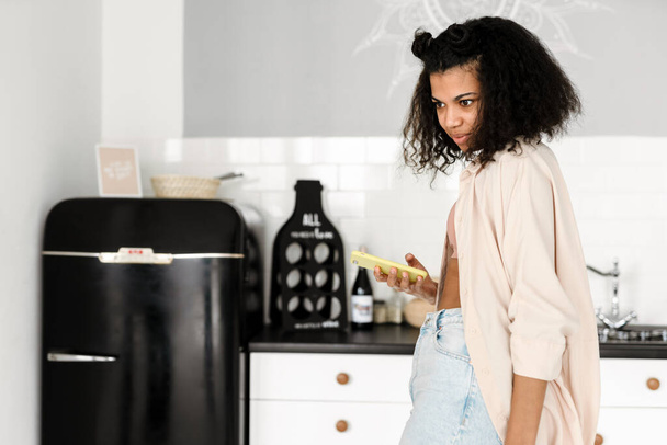 Black young woman using mobile phone in kitchen at home - Фото, зображення