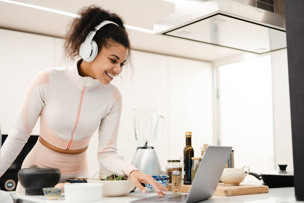 Black woman in headphones using laptop while cooking in kitchen at home - Zdjęcie, obraz