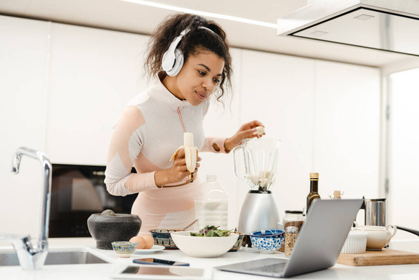 Black woman in headphones using laptop while making smoothie at home - Φωτογραφία, εικόνα