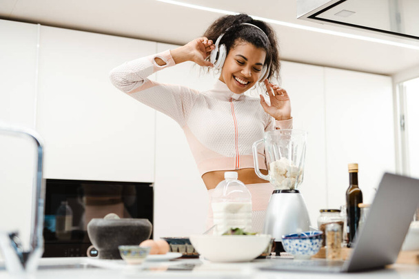 Black woman in headphones using laptop while making smoothie at home - Foto, immagini