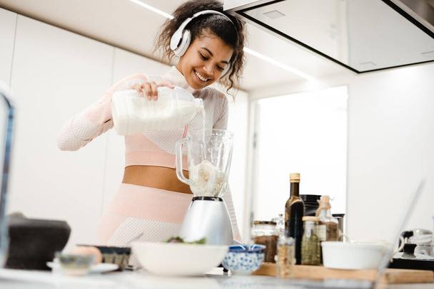 Black woman in headphones smiling and making smoothie with milk at home - Foto, afbeelding