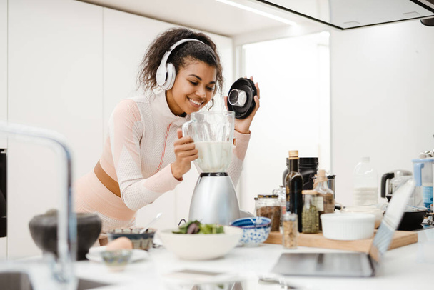 Black woman in headphones smiling and making smoothie in kitchen at home - Foto, Imagen