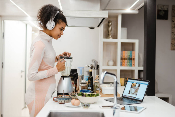 Μαύρη γυναίκα σε ακουστικά χρησιμοποιώντας φορητό υπολογιστή, ενώ κάνοντας smoothie στο σπίτι - Φωτογραφία, εικόνα