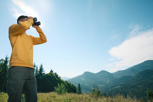 Homem com binóculos nas montanhas no dia ensolarado, vista de baixo ângulo. Espaço para texto - Foto, Imagem