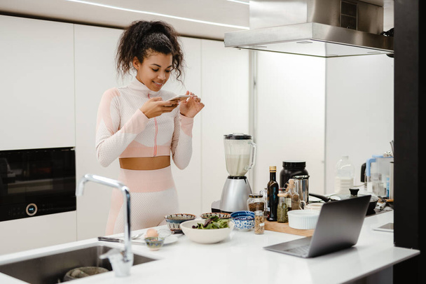 Black woman taking photo on mobile phone while cooking in kitchen at home - Foto, Imagem