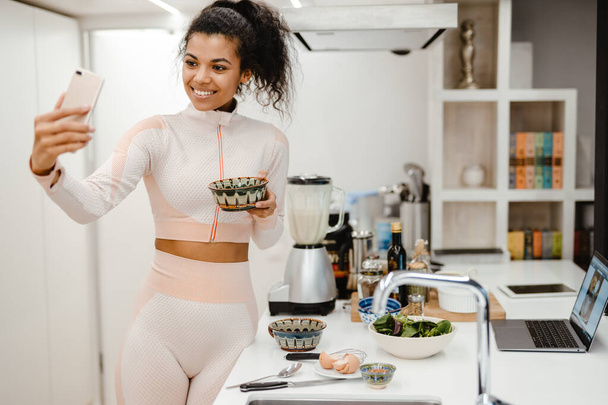 Black woman taking selfie on mobile phone while cooking in kitchen at home - Foto, Imagen